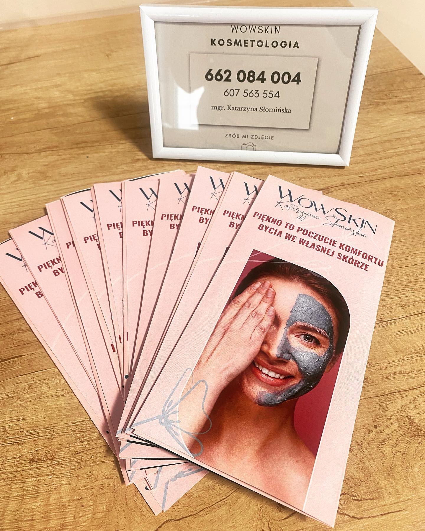 Stos ulotek reklamowych Wowskin Kosmetologia z uśmiechniętą kobietą z maską na połowie twarzy, obok ramka z numerami telefonów na drewnianym blacie.