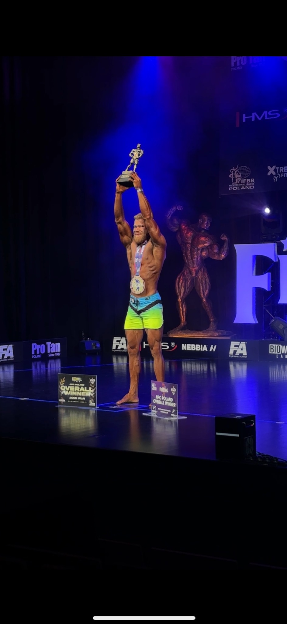 Uśmiechnięty kulturysta z medalem i pucharem na scenie, oświetlony reflektorami, statuetka kulturysty w tle, logo sponsorów. Sukces w zawodach.