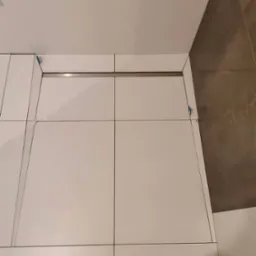 Białe płytki podłogowe w łazience, z widocznymi krzyżykami dystansowymi i odpływem liniowym, częściowo wykończone, z brązową ścianą w tle.