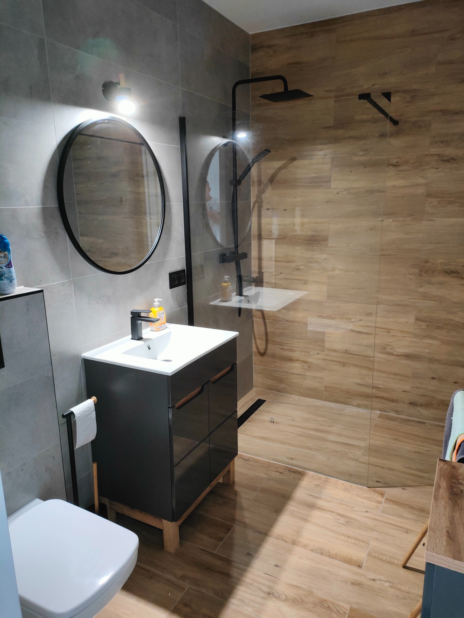 Nowoczesna łazienka z kabiną prysznicową typu walk-in, okrągłym lustrem z czarną ramą i ciemnoszarą szafką pod umywalką z drewnianymi akcentami, wykończona płytkami w odcieniach szarości i drewna.