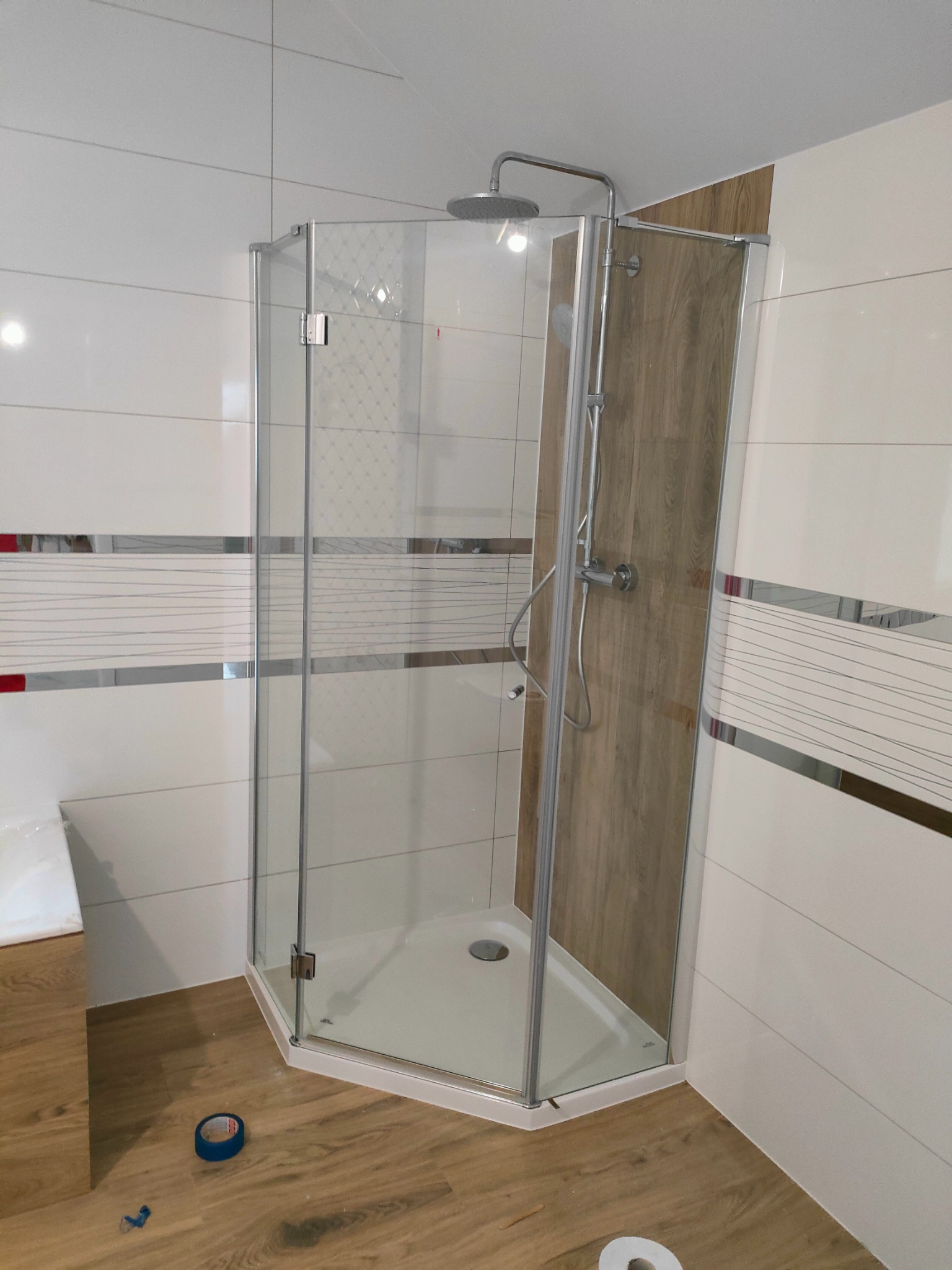 Narożna kabina prysznicowa z chromowanymi profilami, zamontowana w łazience z białymi płytkami ozdobionymi poziomymi paskami i drewnopodobną ścianką, widoczna taśma malarska na podłodze.