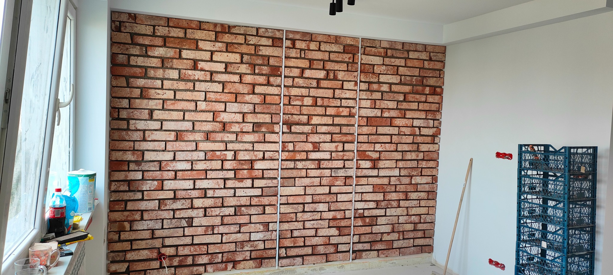 Ściana wykończona czerwoną cegłą klinkierową, podzielona białymi listwami, widoczne puszki instalacyjne, okno z napojami i narzędzia w skrzynce.