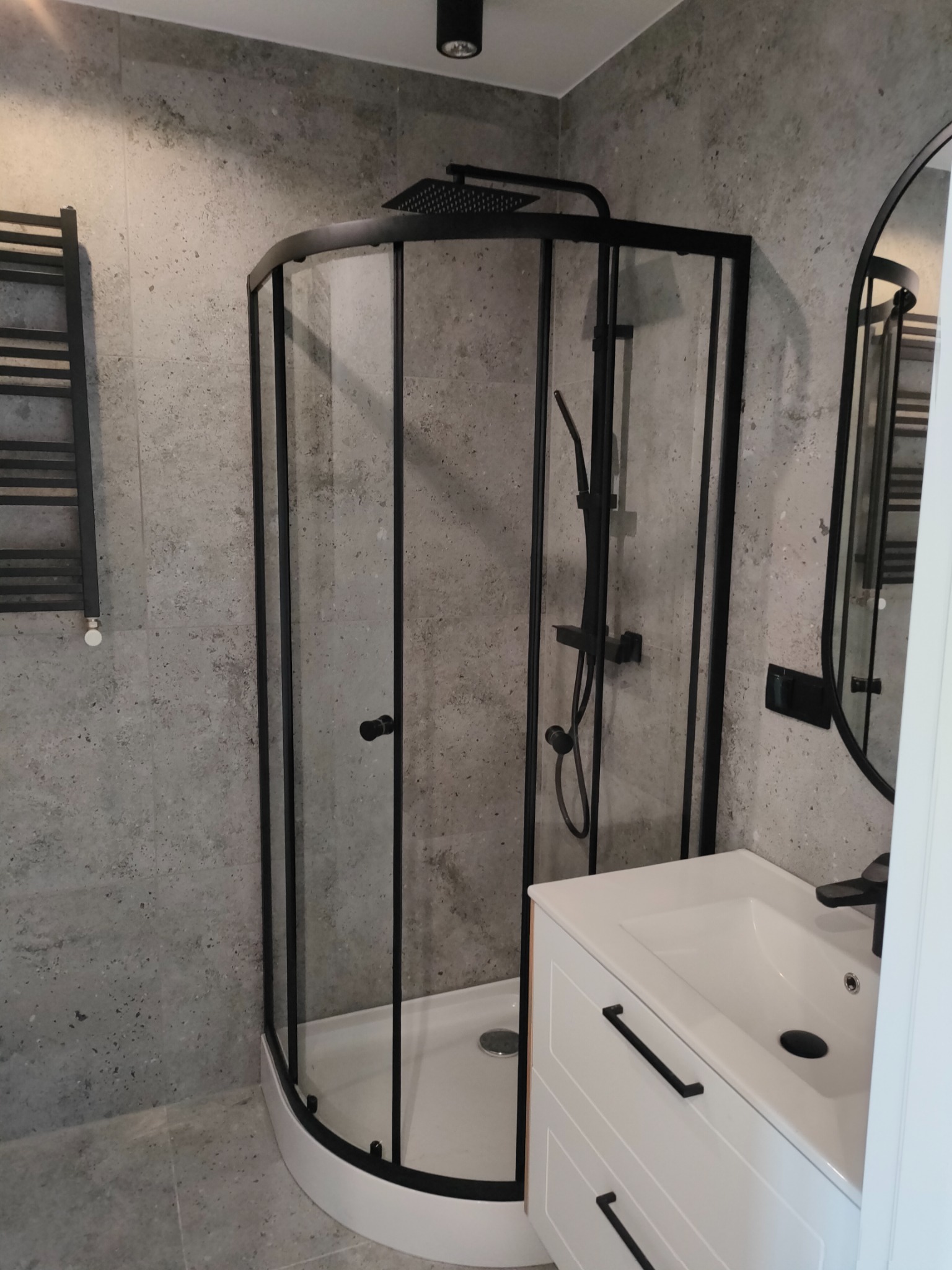 Nowoczesna łazienka z kabiną prysznicową narożną z czarnymi profilami, grzejnikiem drabinkowym i umywalką na szafce z czarnymi uchwytami na tle szarej ściany imitującej beton.