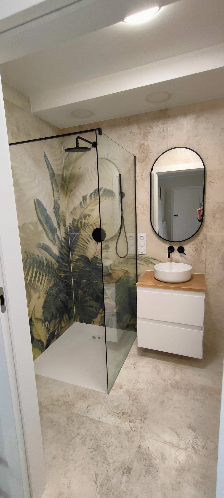 Nowoczesna łazienka z kabiną prysznicową typu walk-in z motywem tropikalnych liści, umywalką nablatową na szafce z drewnianym blatem i owalnym lustrem w czarnej ramie.