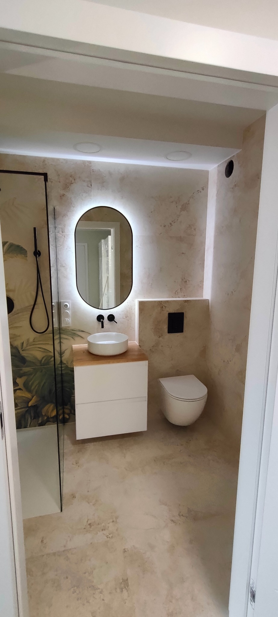 Nowoczesna łazienka z beżowymi ścianami imitującymi kamień, owalne lustro z podświetleniem LED, biała umywalka nablatowa na drewnianym blacie i podwieszana toaleta.