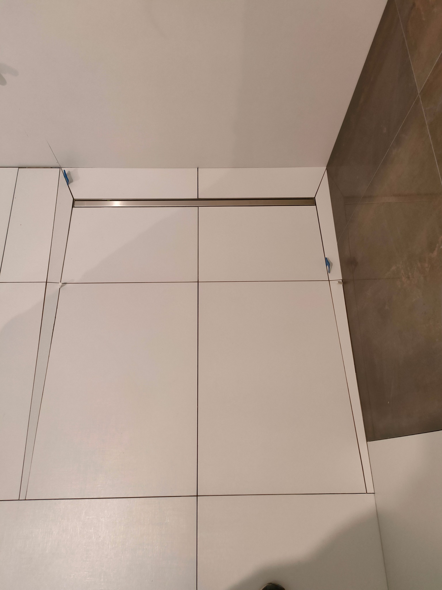 Białe płytki podłogowe w łazience, z widocznymi krzyżykami dystansowymi i odpływem liniowym, częściowo wykończone, z brązową ścianą w tle.