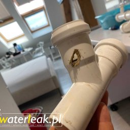WaterLeak.pl - Osuszanie Budynków Wykrywanie Wycieków Wody Wynajem Osuszaczy Lokalizacja Przecieków Gdynia Gdańsk Sopot Warszawa - Uszkodzony, biały plastikowy syfon z widoczną dziurą i pozostałościami po wycieku, trzymany w dłoni, w tle fragment łazienki z oknami dachowymi.