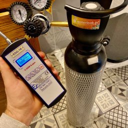 WaterLeak.pl - Osuszanie Budynków Wykrywanie Wycieków Wody Wynajem Osuszaczy Lokalizacja Przecieków Gdynia Gdańsk Sopot Warszawa - Sprawdzanie szczelności instalacji gazowej: detektor gazu EDG-7K w dłoni, wskazujący 0.0% DGW, podłączony do reduktora i manometrów na butli z gazem, tło stanowią wzorzyste płytki podłogowe.