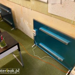 WaterLeak.pl - Osuszanie Budynków Wykrywanie Wycieków Wody Wynajem Osuszaczy Lokalizacja Przecieków Gdynia Gdańsk Sopot Warszawa - Dwa niebieskie panele grzewcze na nóżkach stojące w rogu pokoju z zieloną wykładziną, obok biurko z przyborami do pisania i książkami, oraz biały grzejnik, na ścianie widoczne ślady po zalaniu...