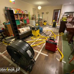 WaterLeak.pl - Osuszanie Budynków Wykrywanie Wycieków Wody Wynajem Osuszaczy Lokalizacja Przecieków Gdynia Gdańsk Sopot Warszawa - Pokój po zalaniu z rozłożonym sprzętem do osuszania: wentylator, osuszacz i węże na podłodze. Widoczne ślady po usunięciu paneli podłogowych i logo firmy na pierwszym planie.