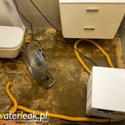 WaterLeak.pl - Osuszanie Budynków Wykrywanie Wycieków Wody Wynajem Osuszaczy Lokalizacja Przecieków Gdynia Gdańsk Sopot Warszawa - Osuszanie posadzki w łazience po zalaniu: biała toaleta podwieszana, biała szafka z szufladami, wentylator podłogowy i osuszacz kondensacyjny z żółtymi wężami. Widoczne ślady wilgoci na podłodze.