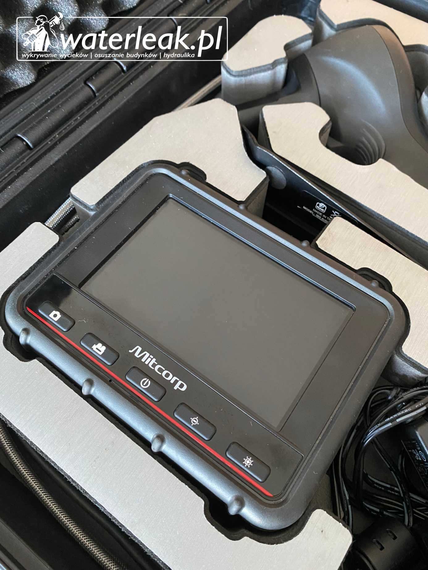 Urządzenie Mitcorp z ekranem i przyciskami funkcyjnymi, w sztywnym futerale, używane do wykrywania wycieków i inspekcji hydraulicznych.