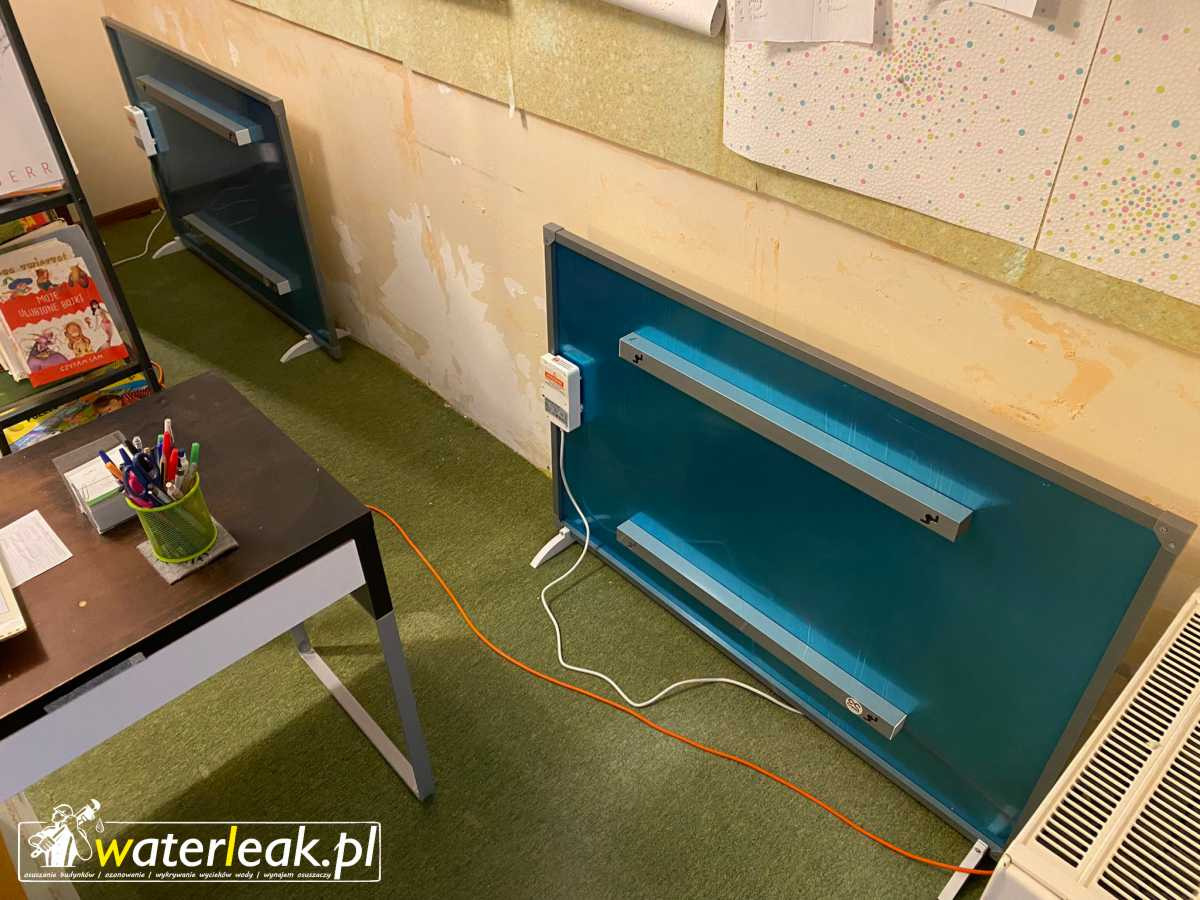 Dwa niebieskie panele grzewcze na nóżkach stojące w rogu pokoju z zieloną wykładziną, obok biurko z przyborami do pisania i książkami, oraz biały grzejnik, na ścianie widoczne ślady po zalaniu...