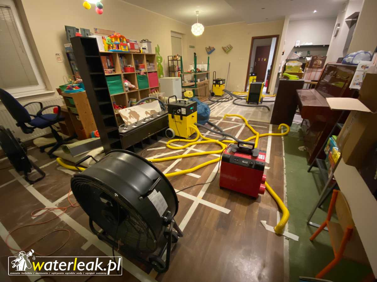 Pokój po zalaniu z rozłożonym sprzętem do osuszania: wentylator, osuszacz i węże na podłodze. Widoczne ślady po usunięciu paneli podłogowych i logo firmy na pierwszym planie.