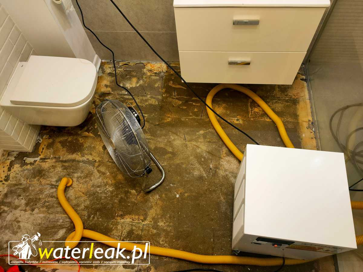Osuszanie posadzki w łazience po zalaniu: biała toaleta podwieszana, biała szafka z szufladami, wentylator podłogowy i osuszacz kondensacyjny z żółtymi wężami. Widoczne ślady wilgoci na podłodze.