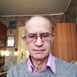 Portret mężczyzny w okularach, z widocznym fragmentem pokoju z boazerią i meblami w tle.