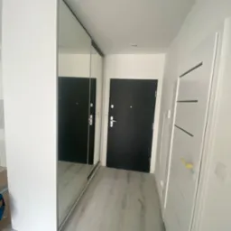 Biała szafa z lustrzanymi drzwiami przesuwnymi w przedpokoju, widoczne drzwi wejściowe i drzwi wewnętrzne z poziomymi zdobieniami, szare panele na podłodze.