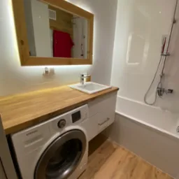 Nowoczesna łazienka z drewnianym blatem nad pralką, lustrem z podświetleniem LED w drewnianej ramie i drewnianą podłogą.