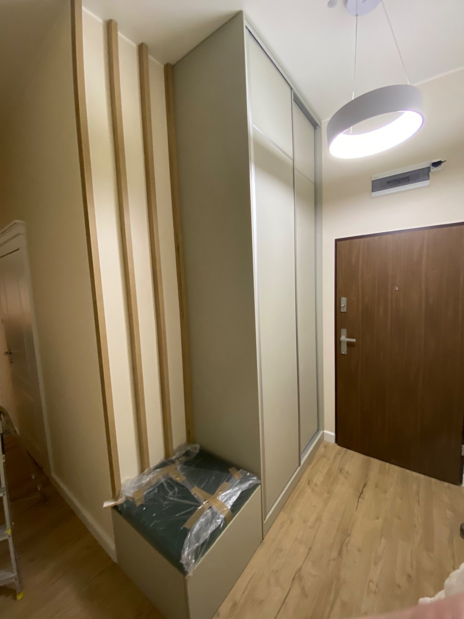 Korytarz z szafą przesuwną w zabudowie, dekoracyjnymi listwami pionowymi na ścianie, siedziskiem w folii ochronnej i nowoczesną lampą sufitową