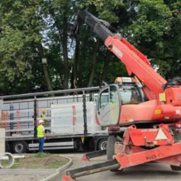Rozładunek metalowych elementów konstrukcyjnych z naczepy ciężarówki przy użyciu czerwonego wózka widłowego Manitou, pracownicy w kamizelkach odblaskowych nadzorują proces.