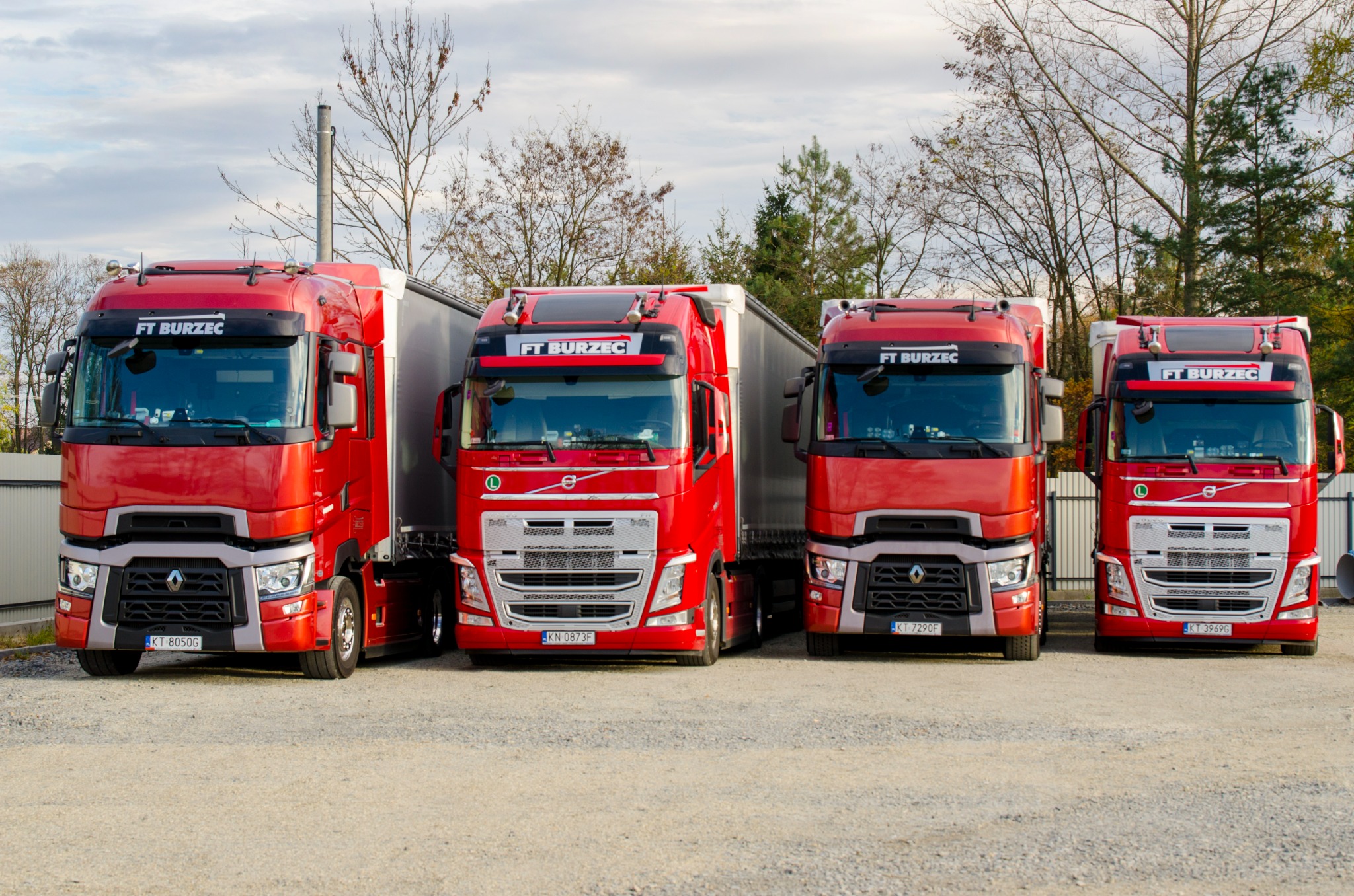 Cztery czerwone ciężarówki z naczepami, marki Renault i Volvo, zaparkowane obok siebie z widocznym logo firmy 'FT BURZEC' na przedniej szybie, na żwirowym parkingu.