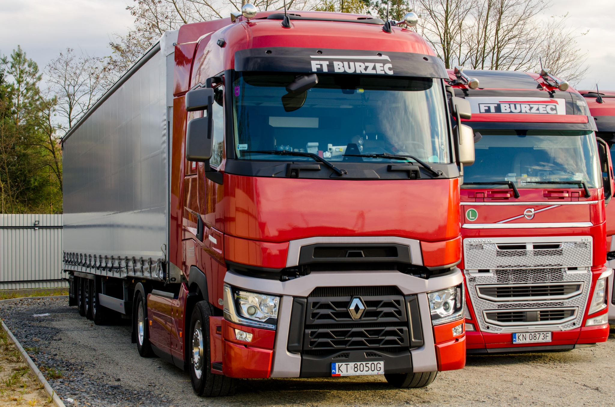 Dwa czerwone ciągniki siodłowe, Renault i Volvo, stojące obok siebie na żwirowym parkingu. Na pierwszym planie ciągnik Renault z naczepą, widoczny numer rejestracyjny KT 8050G.