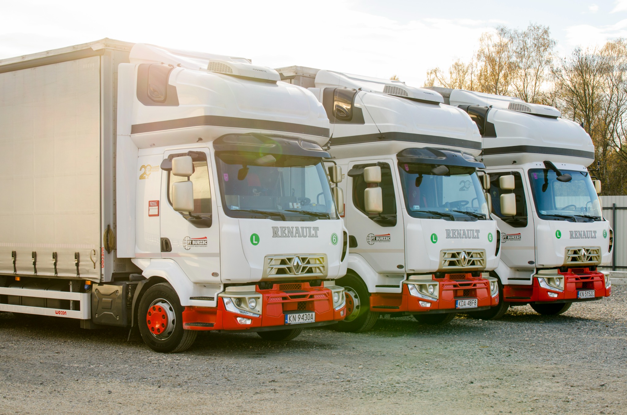 Trzy białe ciężarówki Renault z czerwonymi akcentami zaparkowane na żwirowym placu, widoczne logo firmy transportowej FT Burzec i oznaczenia 'L' oraz numery rejestracyjne.
