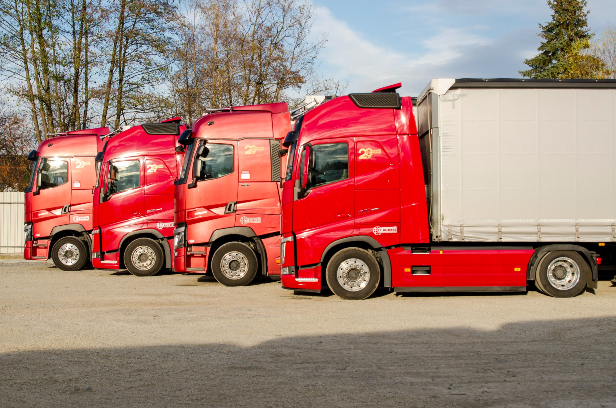 Czerwone ciągniki siodłowe z naczepami, ustawione w rzędzie na parkingu, z logo firmy FT BURZEC na kabinach.