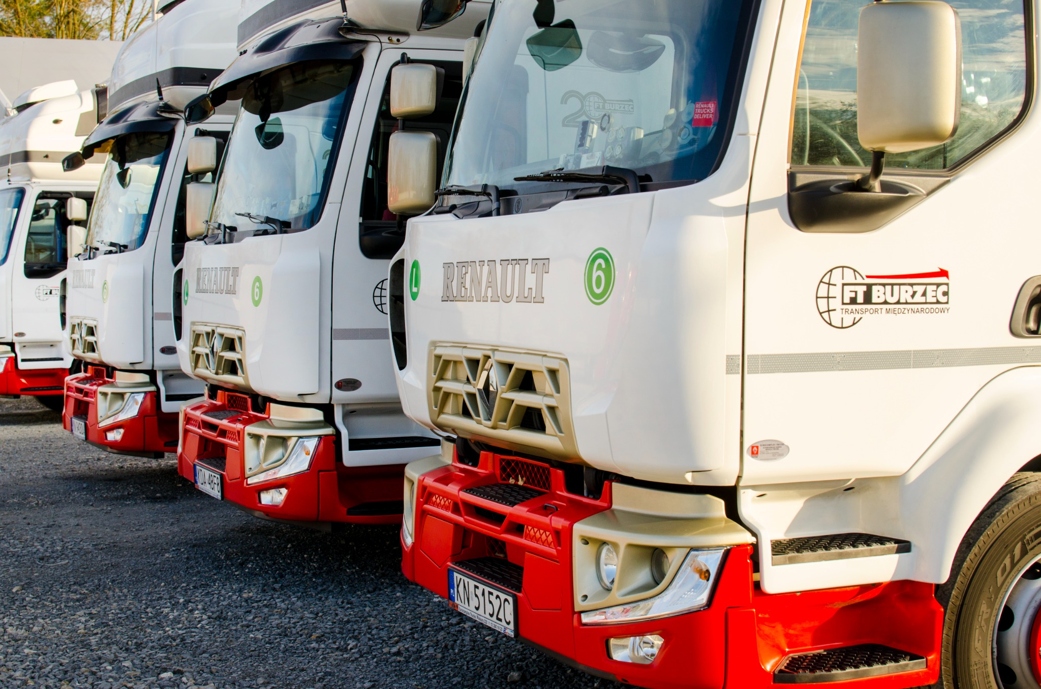 Flota białych ciężarówek Renault firmy FT Burzec zaparkowana na żwirowym placu, z widocznym logo firmy na boku kabiny i czerwonymi elementami nadwozia.