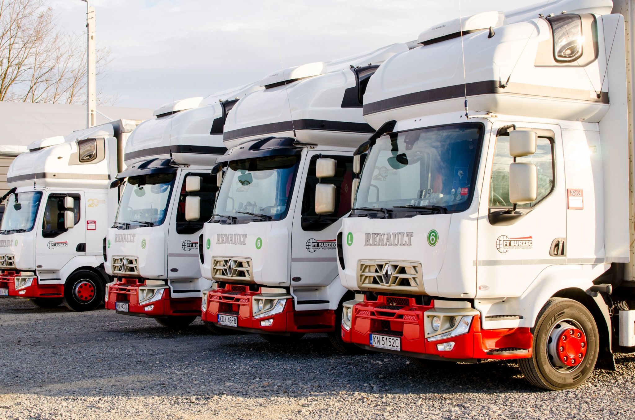 Szereg białych ciężarówek Renault z czerwonymi akcentami, zaparkowanych na żwirowym parkingu, z logo firmy FT Burzec na boku kabiny.
