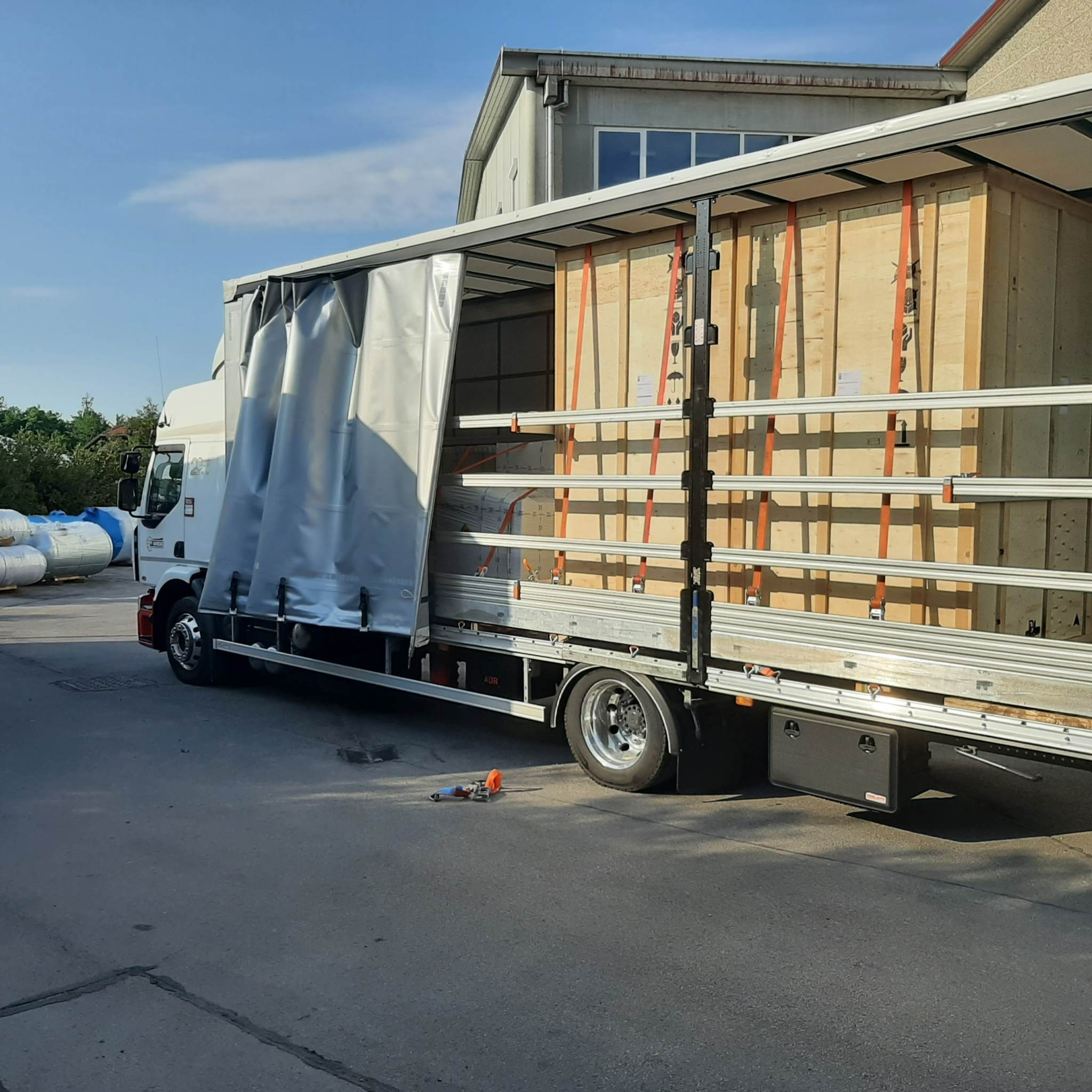 Ciężarówka z częściowo odsłoniętą plandeką, przewożąca dużą, drewnianą skrzynię zabezpieczoną pomarańczowymi pasami transportowymi.