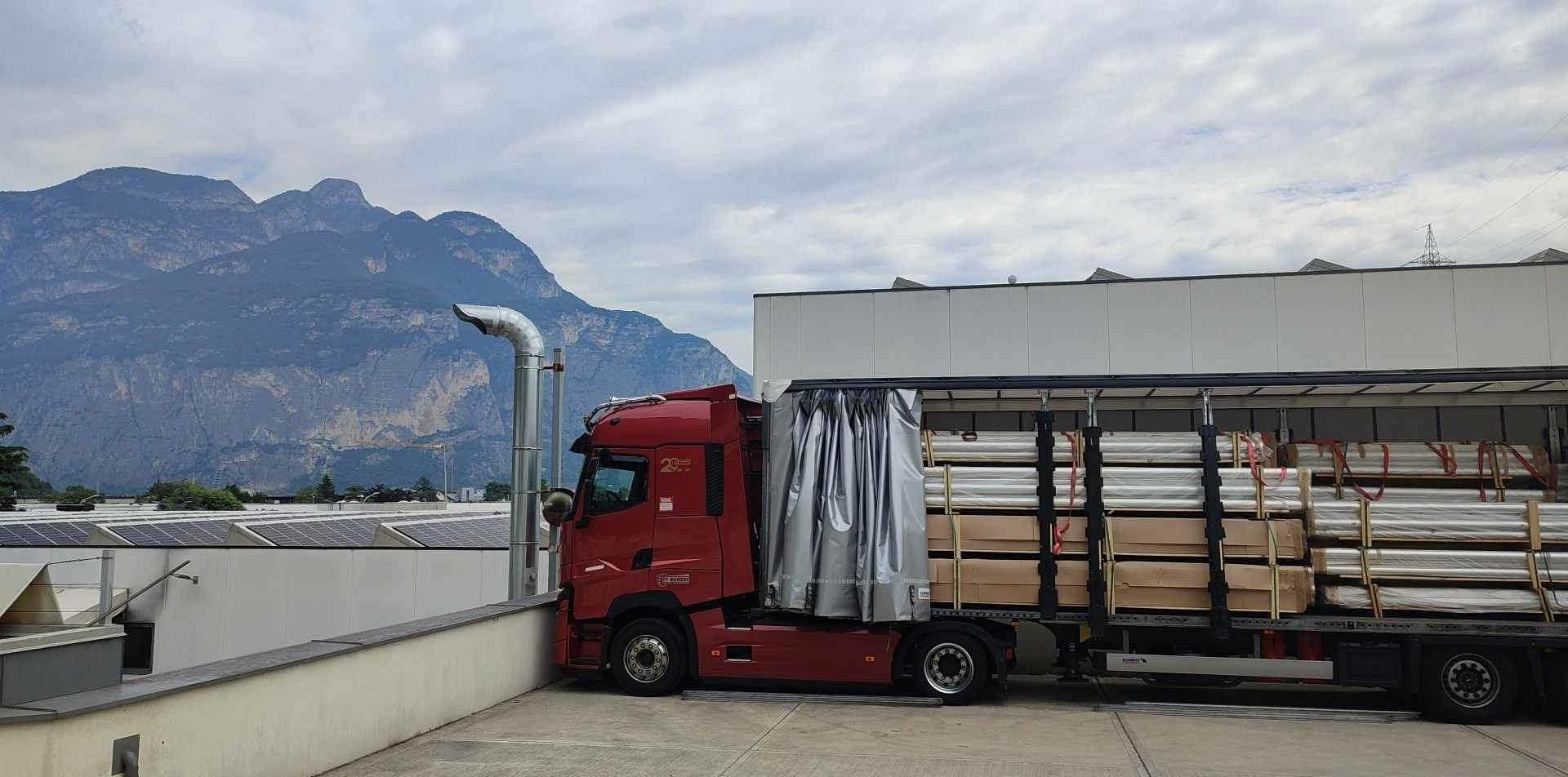Czerwona ciężarówka z naczepą załadowana metalowymi rurami i kartonami, na tle górskiego krajobrazu i budynku z panelami słonecznymi na dachu.