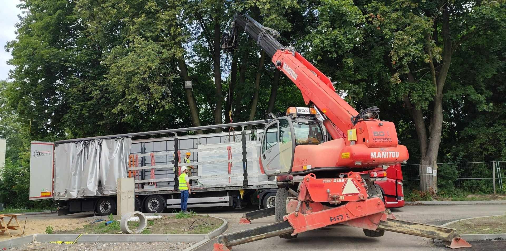 Rozładunek metalowych elementów konstrukcyjnych z naczepy ciężarówki przy użyciu czerwonego wózka widłowego Manitou, pracownicy w kamizelkach odblaskowych nadzorują proces.
