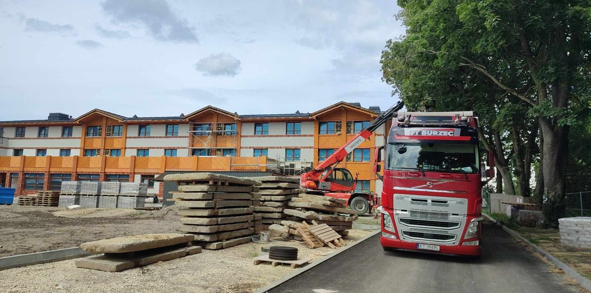 Czerwona ciężarówka firmy transportowej z napisem FT BURZEC na tle budynku w trakcie budowy i sterty kamiennych płyt.