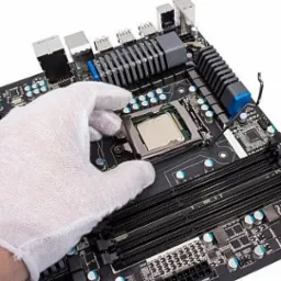 Biała rękawica umieszcza procesor w gnieździe czarnej płyty głównej z widocznymi slotami pamięci RAM i radiatorem.