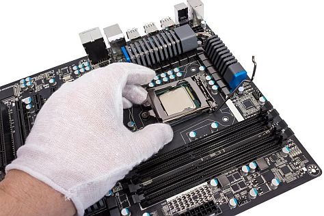 Biała rękawica umieszcza procesor w gnieździe czarnej płyty głównej z widocznymi slotami pamięci RAM i radiatorem.