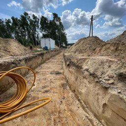 Mateo Mateusz Klikowski - Głęboki, wykopany rów w piaszczystej ziemi, z żółtym kablem zwiniętym w kłębek na pierwszym planie, białą przyczepą i koparką w tle, pod błękitnym niebem z obłokami.
