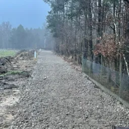 Utwardzona droga szutrowa biegnąca przez las w pochmurny dzień, widoczne białe słupki wyznaczające trasę i ogrodzenie z siatki.