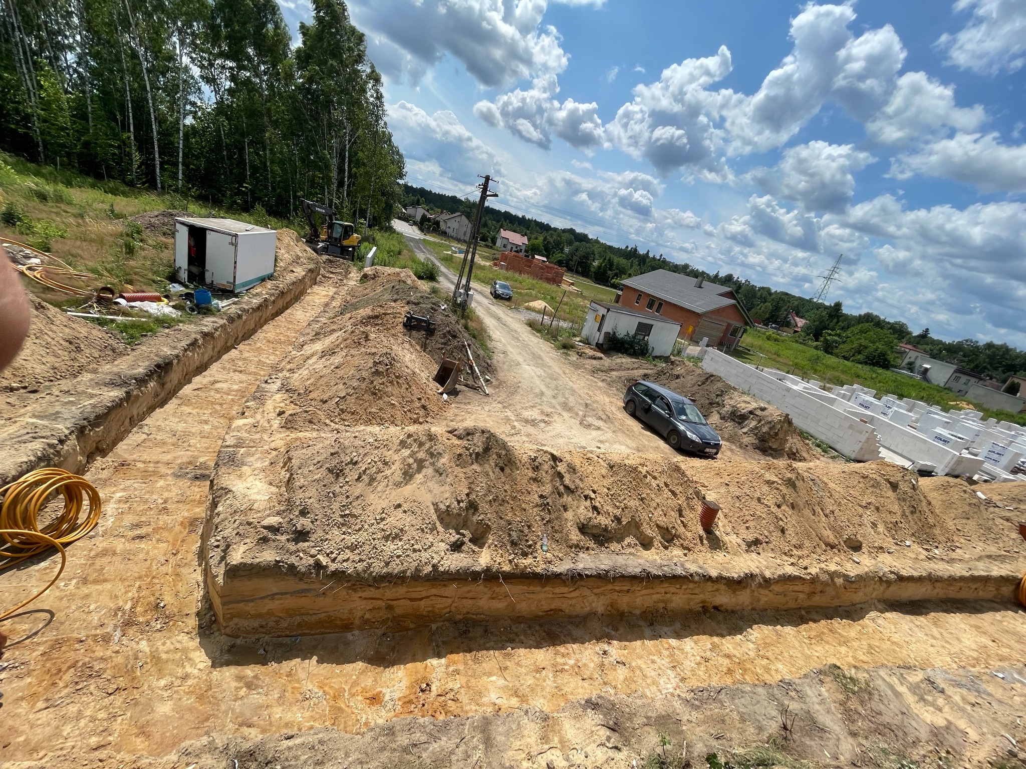 Wykop rowów pod budowę z widocznym sprzętem budowlanym, stertami ziemi i materiałami budowlanymi w tle, pod błękitnym niebem z białymi chmurami.