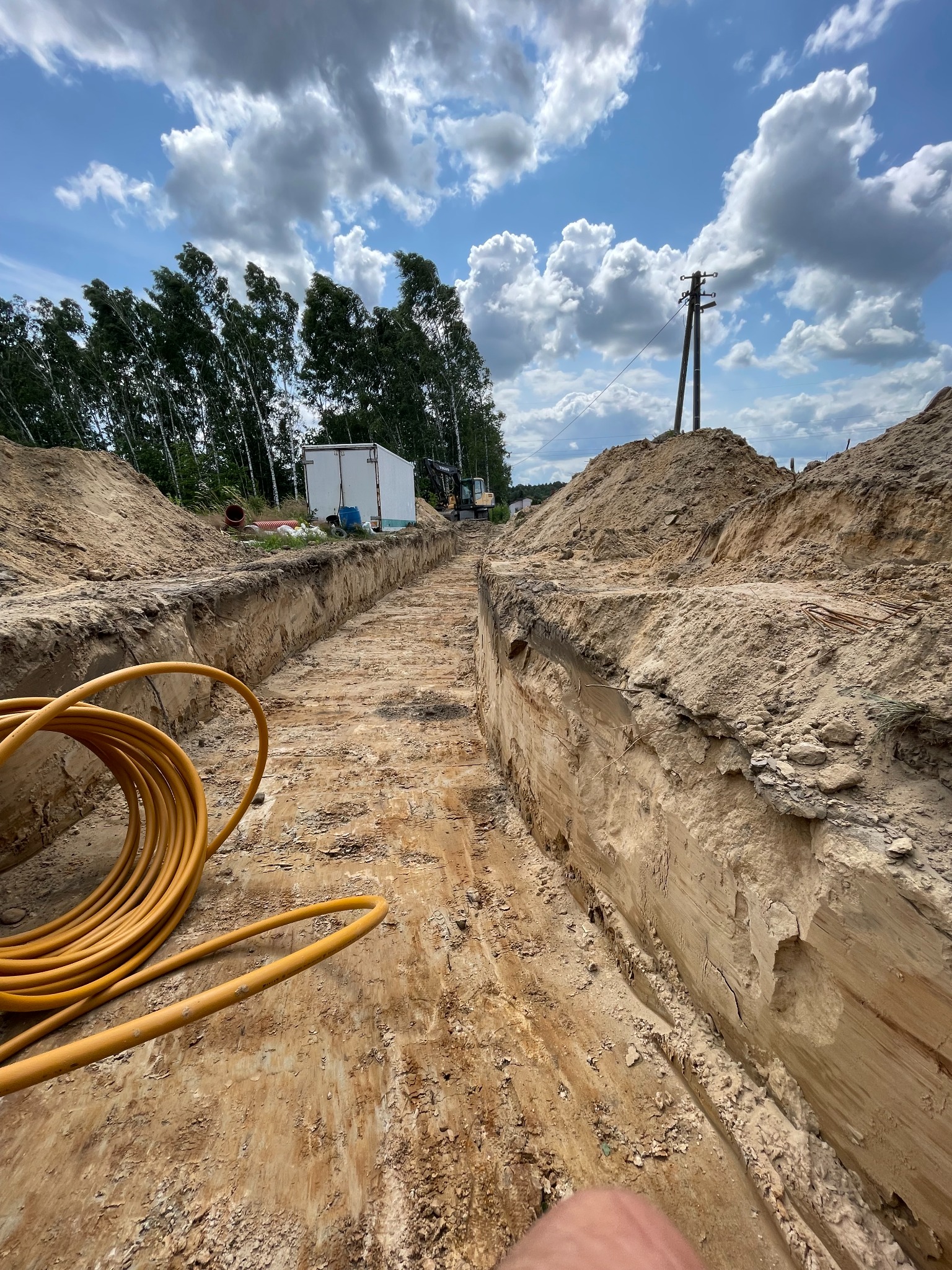 Głęboki, wykopany rów w piaszczystej ziemi, z żółtym kablem zwiniętym w kłębek na pierwszym planie, białą przyczepą i koparką w tle, pod błękitnym niebem z obłokami.