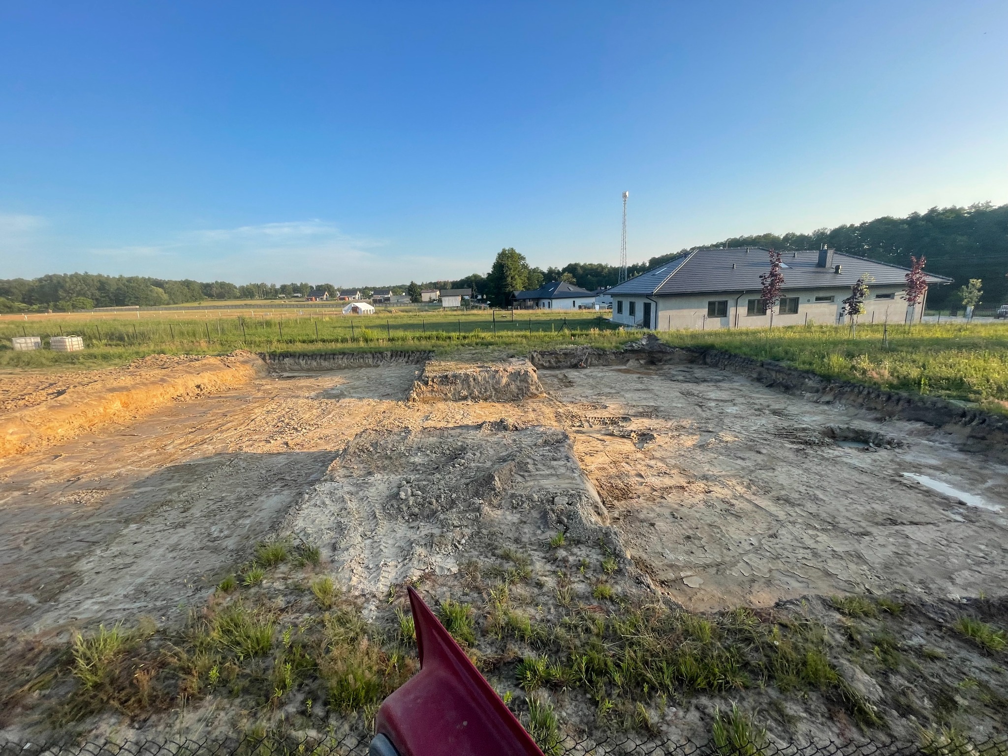 Przygotowany teren pod budowę domu jednorodzinnego z widocznymi wykopami pod fundamenty, zarys budynku w ziemi, w tle dom, łąka i błękitne niebo.