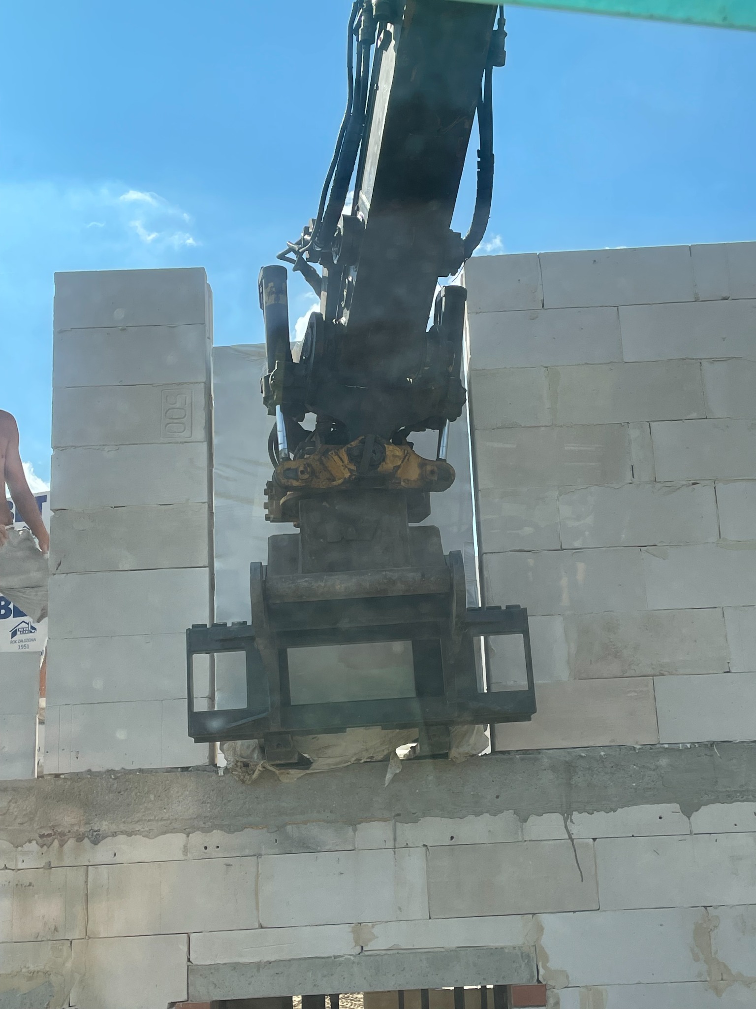 Żuraw przenosi blok betonu komórkowego na budowie ściany, widoczne niebo i fragment pracownika.