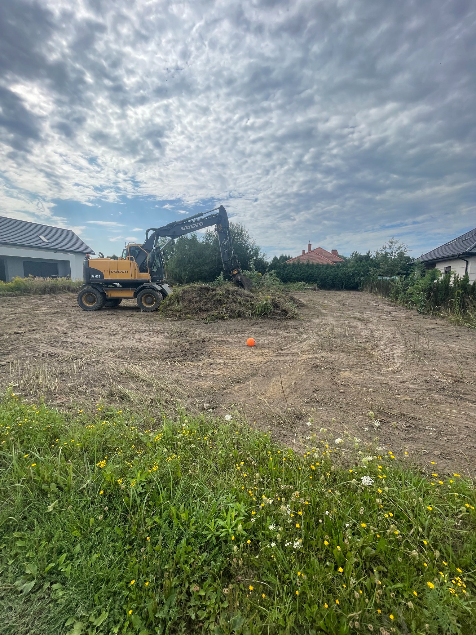 Żółta koparka Volvo EW140D na działce z wykopaną ziemią, widoczne domy w tle i pomarańczowy pachołek.