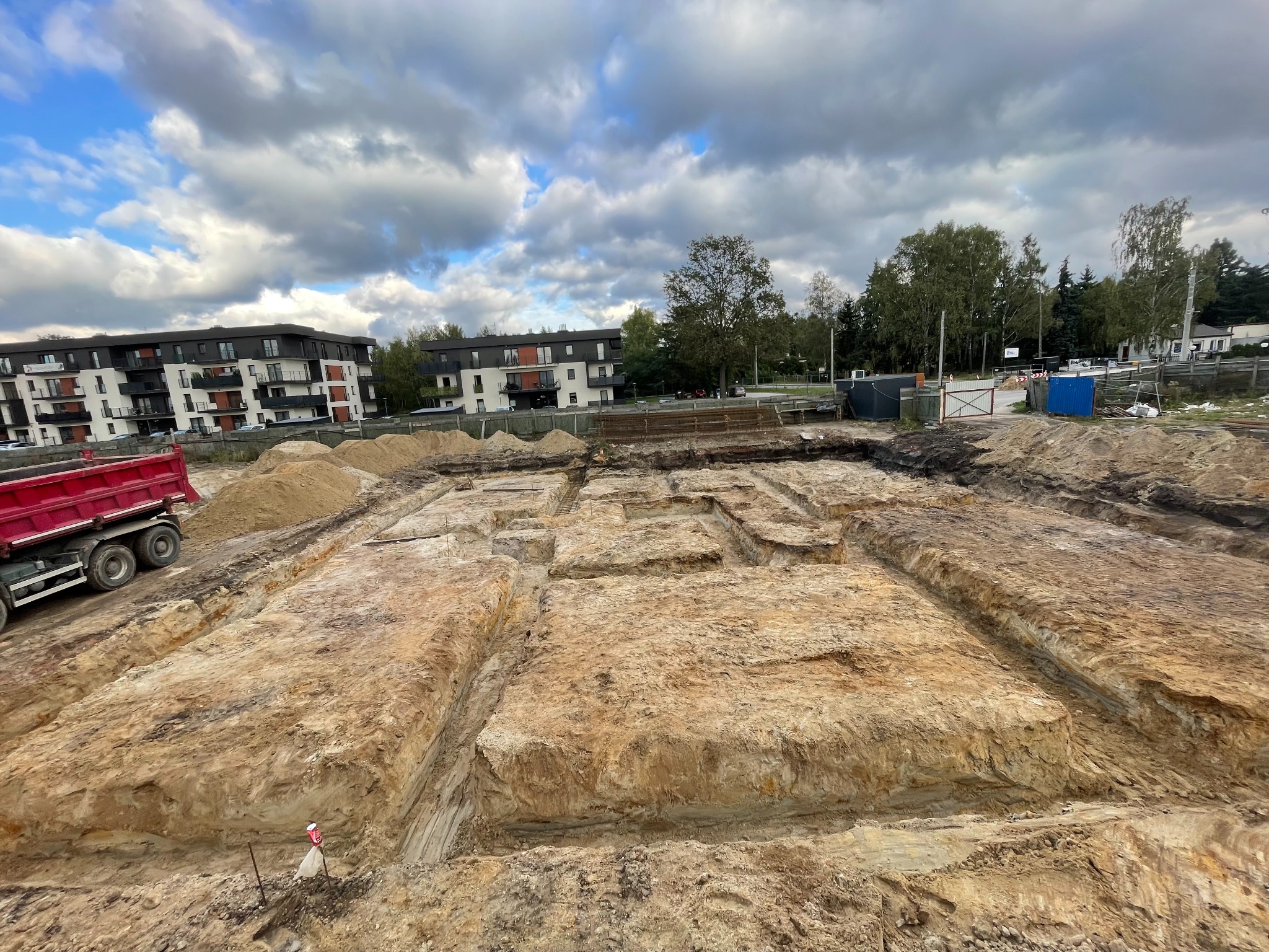 Przygotowany wykop pod fundamenty budynku w Łodzi, widoczny czerwony samochód ciężarowy i budynki w tle, pochmurne niebo.