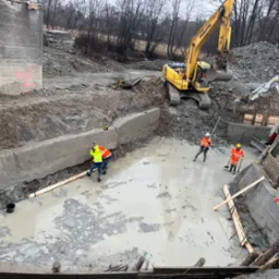 Prace ziemne przy budowie fundamentów w terenie podmokłym, z użyciem koparek i z udziałem pracowników w odblaskowych ubraniach, widoczne drewniane elementy szalunku.