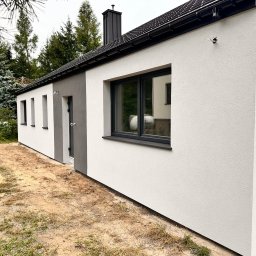 Adam Kaczor - Świeżo otynkowana elewacja domu jednorodzinnego z widocznymi puszkami instalacyjnymi, czarną stolarką okienną i dachem z blachy na rąbek stojący; na pierwszym planie wiaderka po farbach elewacyjnych.