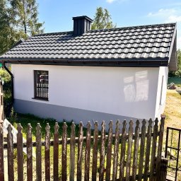 Adam Kaczor - Widok z zewnątrz na otynkowany budynek z nowym dachem z blachodachówki w kolorze grafitowym, z widocznym kominem; w tle drzewa, a na pierwszym planie drewniany płot i stos starych dachówek.