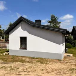 Biały, parterowy budynek z szarym cokołem i czarnym dachem, widok z przodu na tle błękitnego nieba.