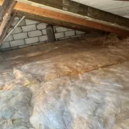 Poddasze z widoczną wełną mineralną pomiędzy drewnianymi belkami stropowymi i fragmentem ściany z bloczków betonowych.