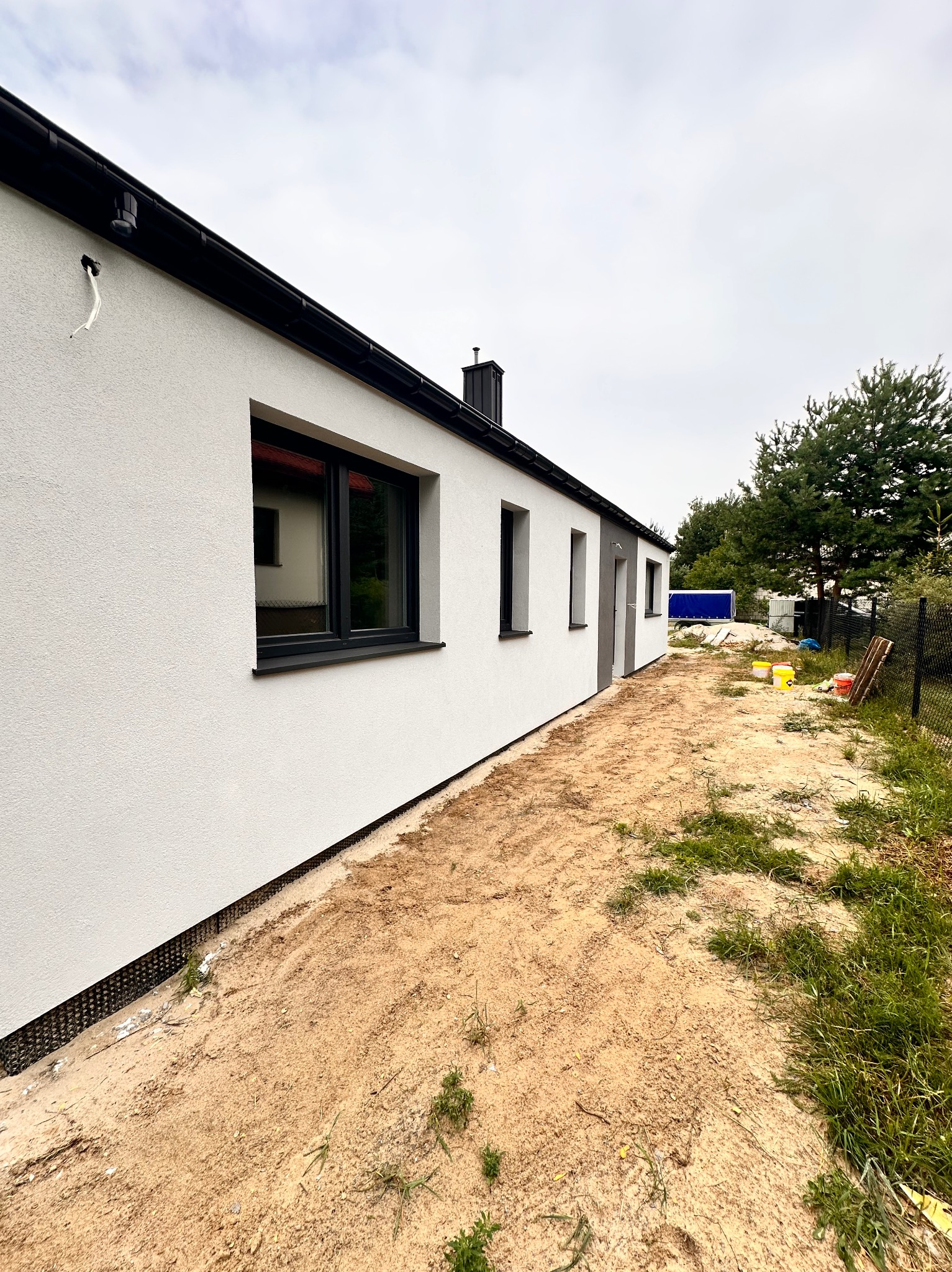 Świeżo otynkowany, biały dom jednorodzinny z czarnymi ramami okiennymi i rynnami, widok z boku na niezagospodarowaną działkę z piaskiem i widocznymi śladami prac budowlanych.